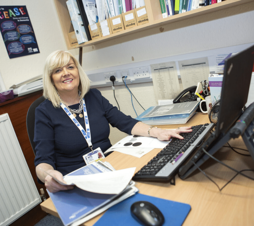 An older blonde lady sat at a busy desk smiling with some paperwork in hand