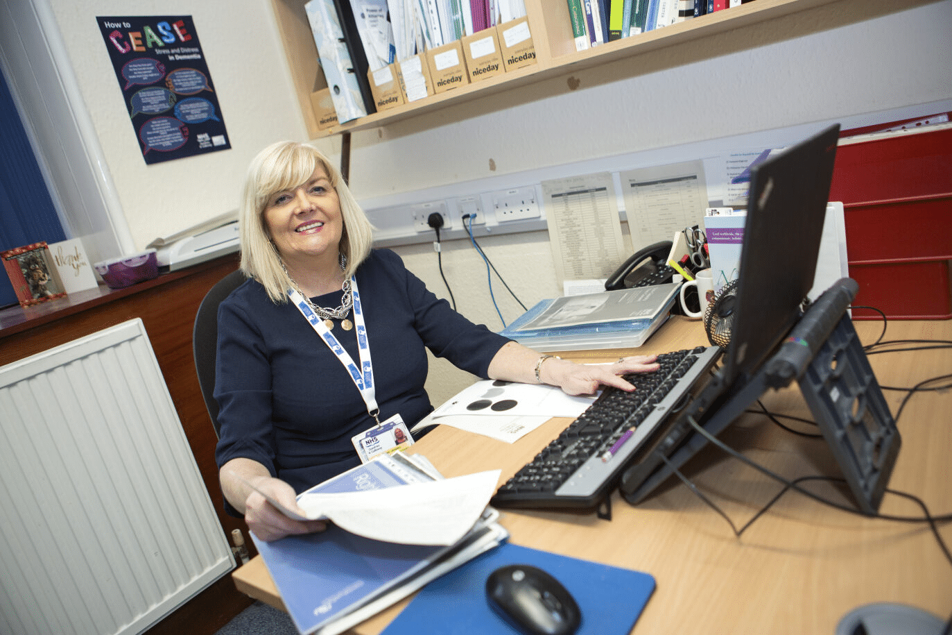An older blonde lady sat at a busy desk smiling with some paperwork in hand