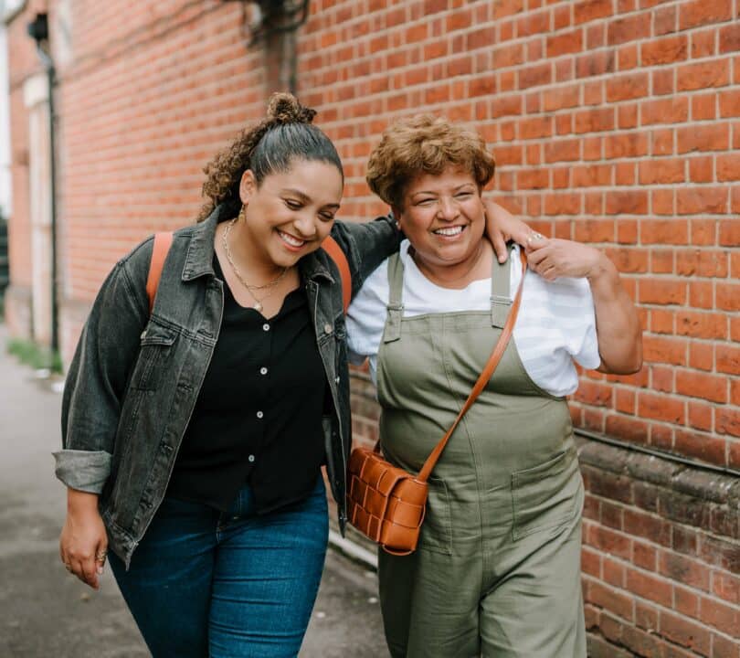 An older and younger woman are walking along outside together. The younger woman has her arm round the older woman. They are both smiling.
