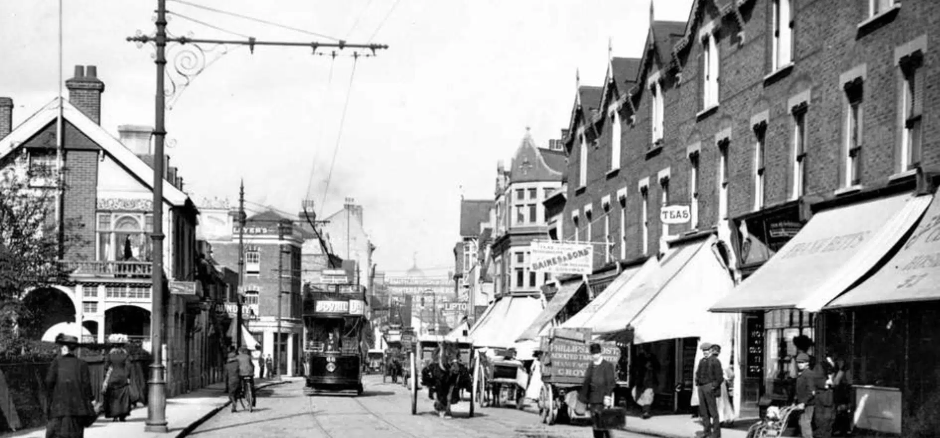 A black and white street scene of a south london street in the 1880s with shopfronts, people and horses
