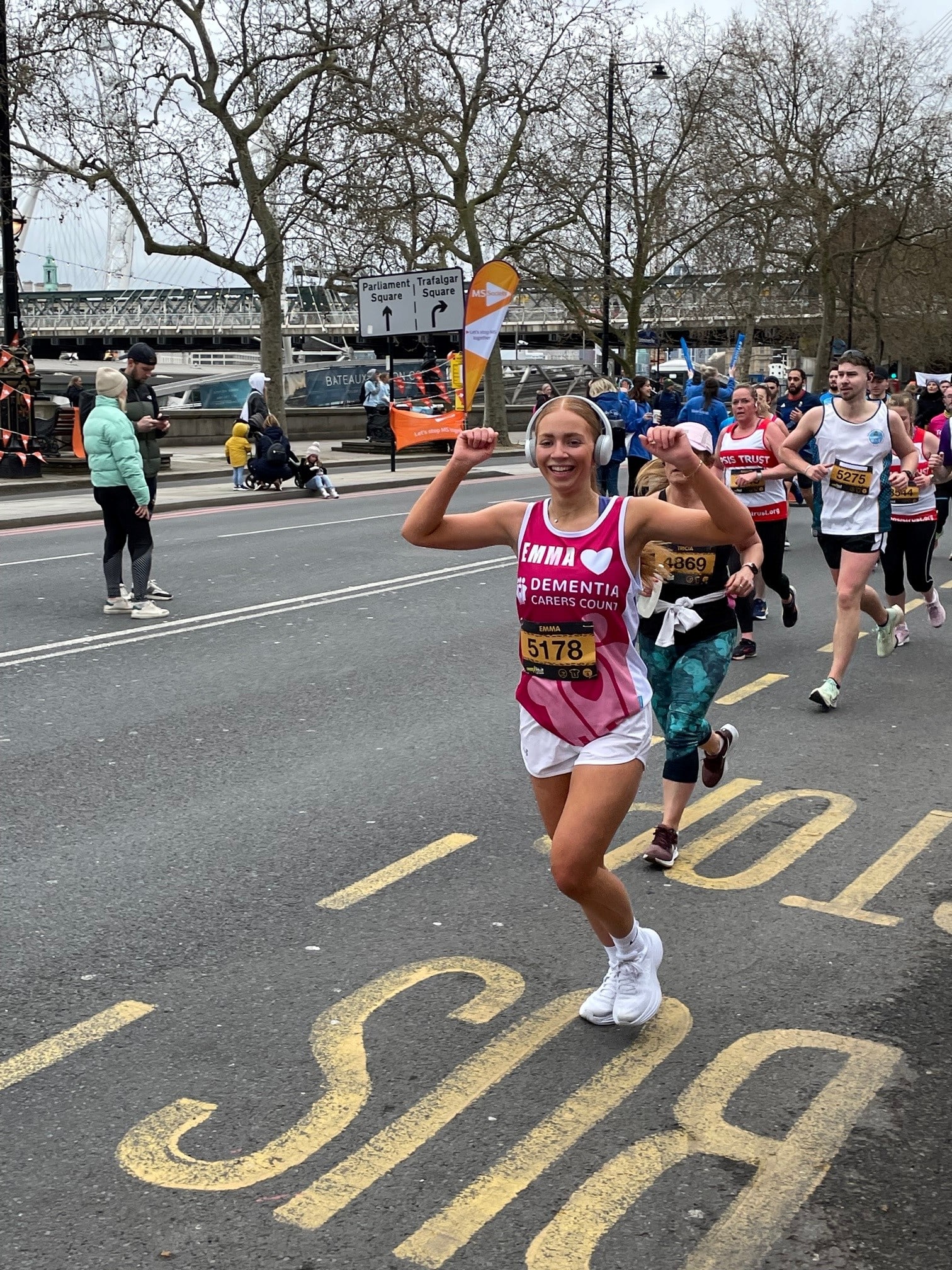 A young woman with long fair hair in a Dementia Carers Count charity running vest raises her arms and smiles at the camera as she runs past during a charity run
