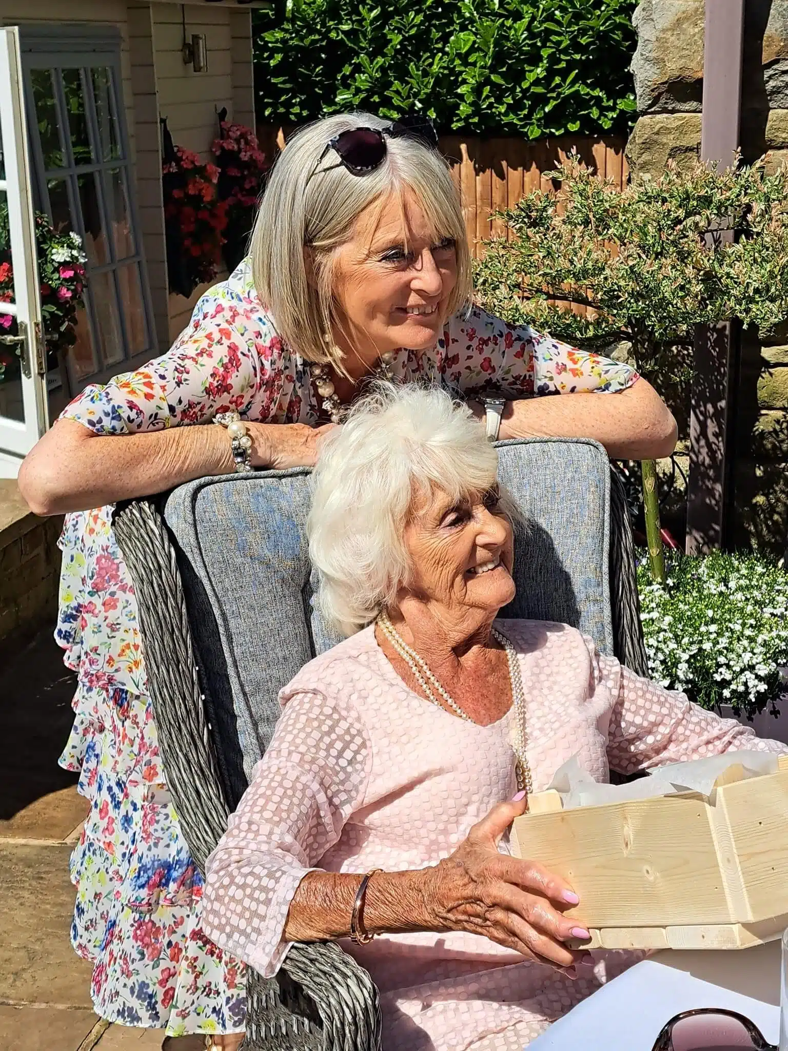 Daughter who struggled to organise care for her mum is outside in the garden smiling and leaning over a chair where her mum is sitting in the sunshine she is also smiling