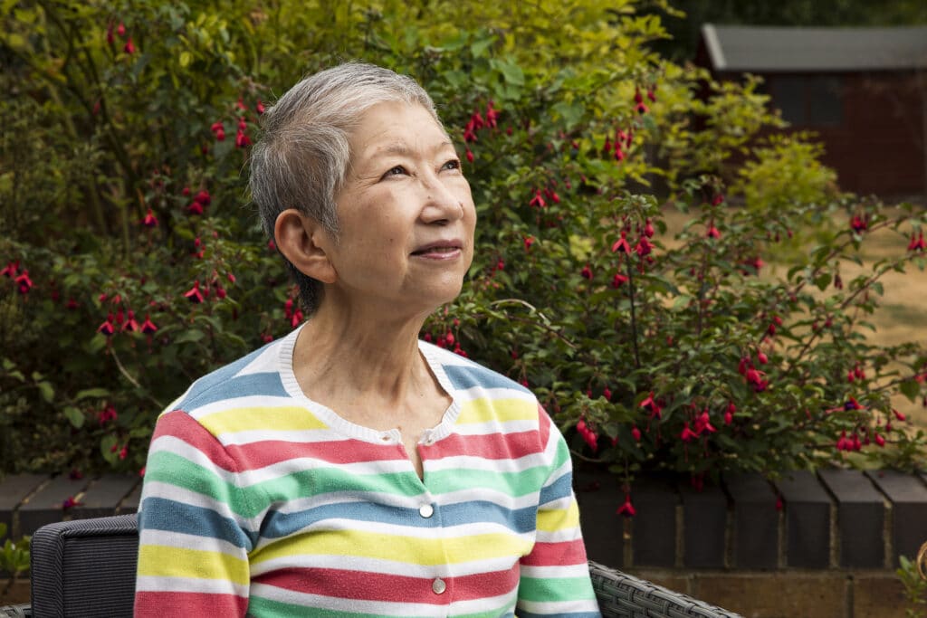 An older Japanese lady with short grey hair and a colourful stripy cardigan looks confidently up, in front of a backdrop of green bushes in her garden.