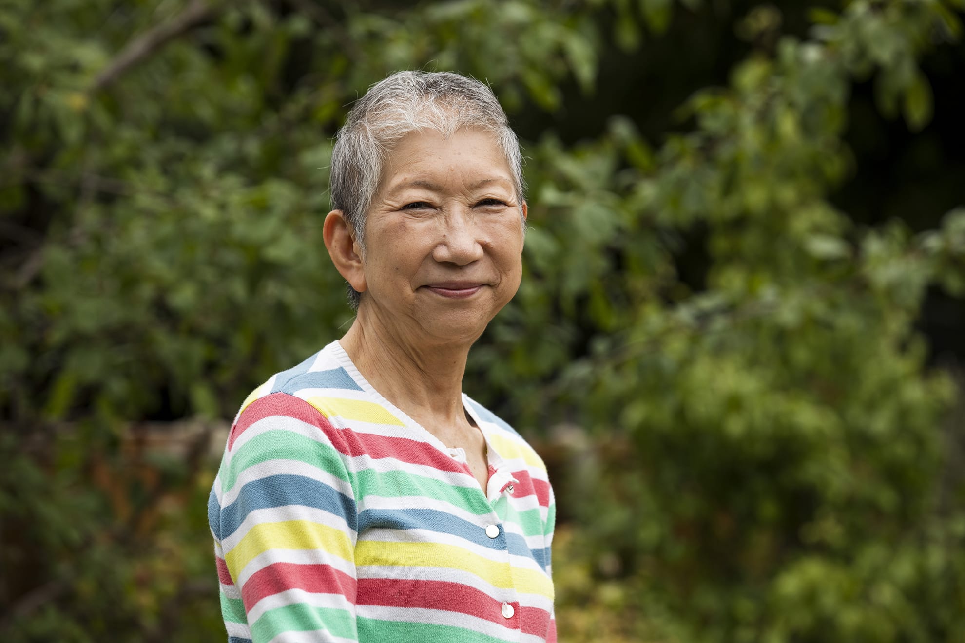An older Japanese lady in a colourful stripy cardigan smiles confidently at the camera in front of some greenery.