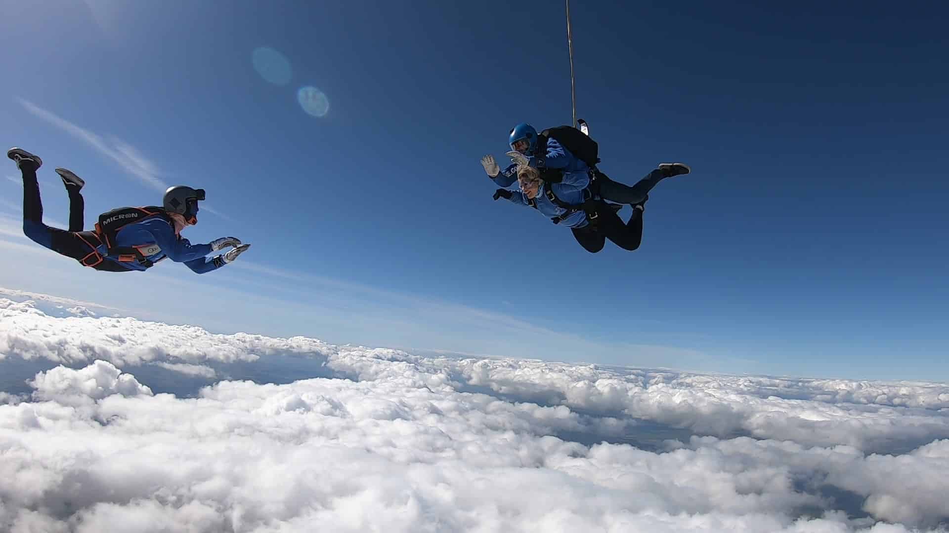 Two figures silhouetted against a blue sky above white clouds, skydiving