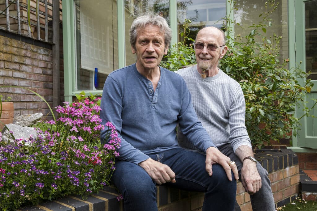 Stuart sits with his partner Roger, who has young onset Alzheimer's, on a low wall in their garden. Stuart and Roger are older men in their 50s and 60s, dressed casually and looking warmly at the camera
