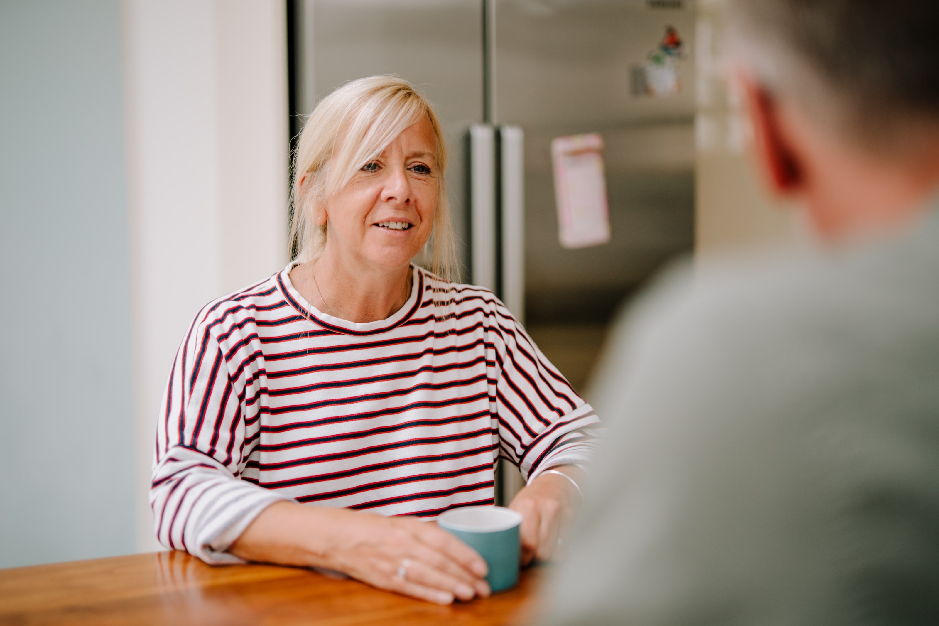 A woman is sitting at a kitchen table talking sensitively to someone while drinking a cup of tea.