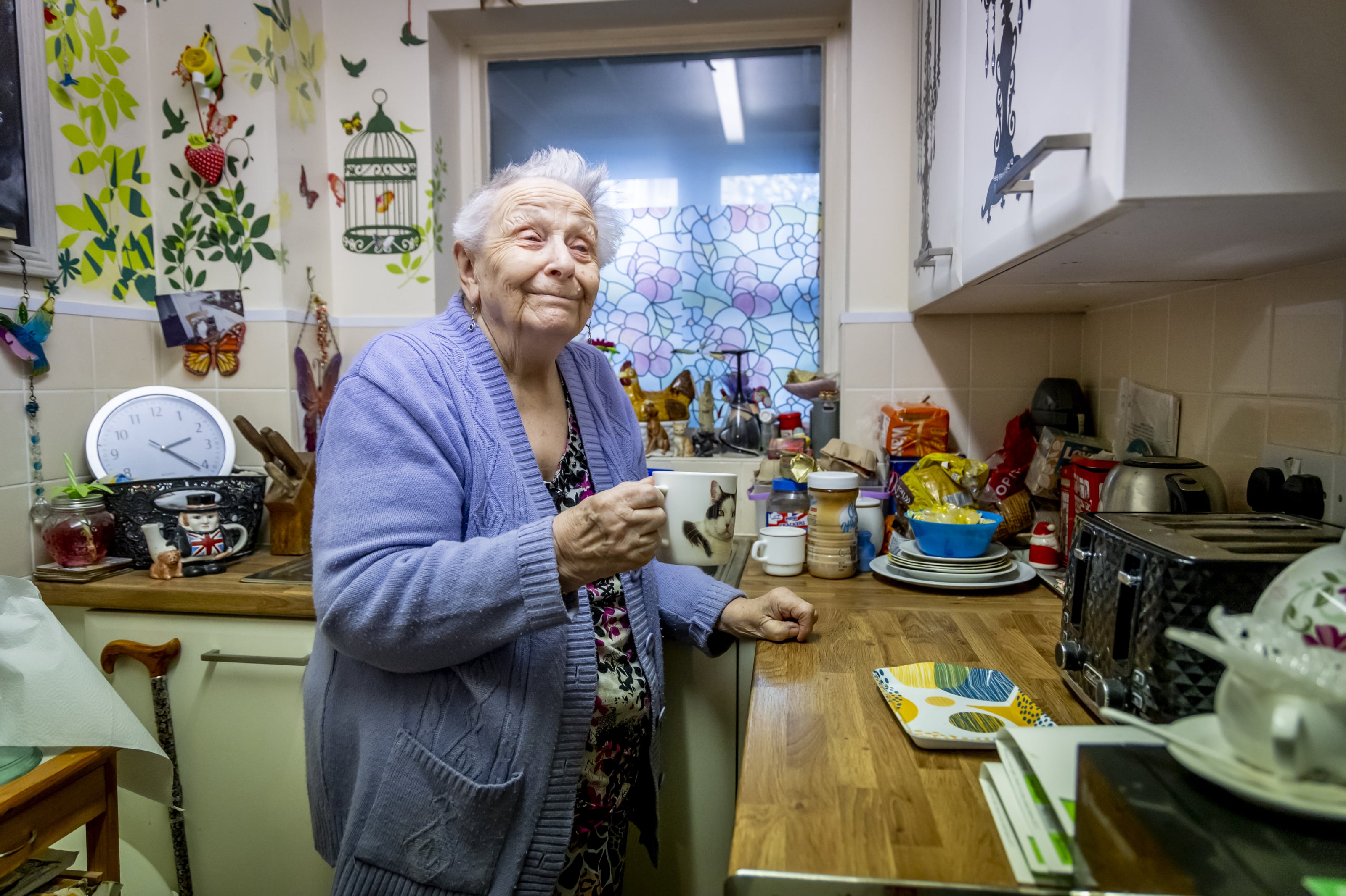 An older lady is in her kitchen and has made a cup of tea. She is smiling.