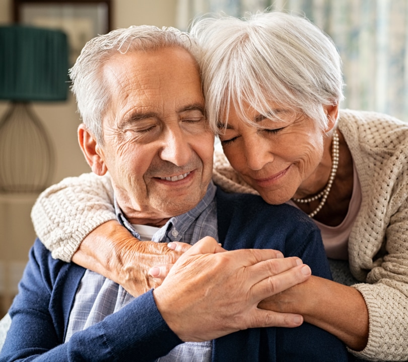 An older couple. The lady wraps her arms around the man's shoulders.