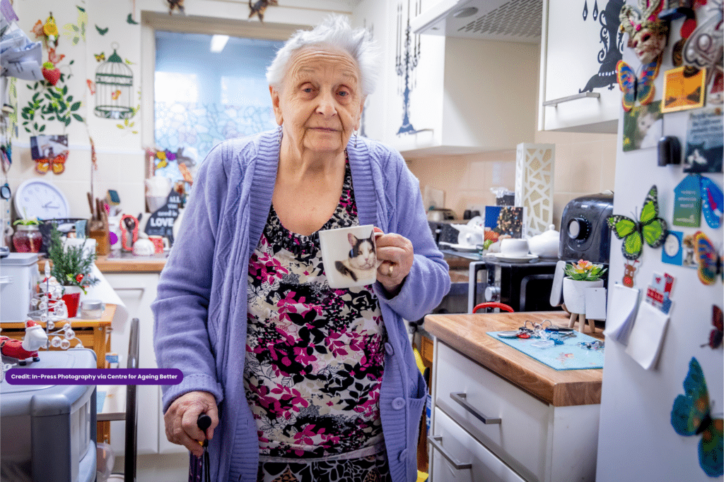 A woman is standing in her kitchen holding a cup of tea and looking at the camera